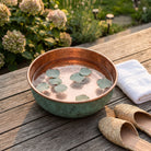 Green patina copper pedicure bowl filled with water on a timber deck beside a garden