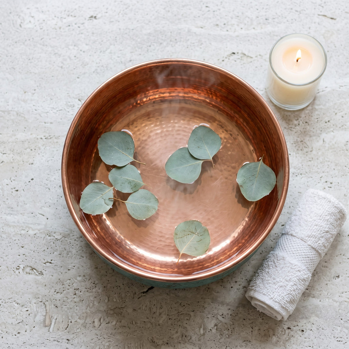 Copper pedicure bowl with eucalyptus, a lit candle and a rolled white towel