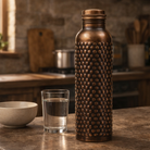 A pure copper water bottle with antique embossing placed on a stone surface in a kitchen.